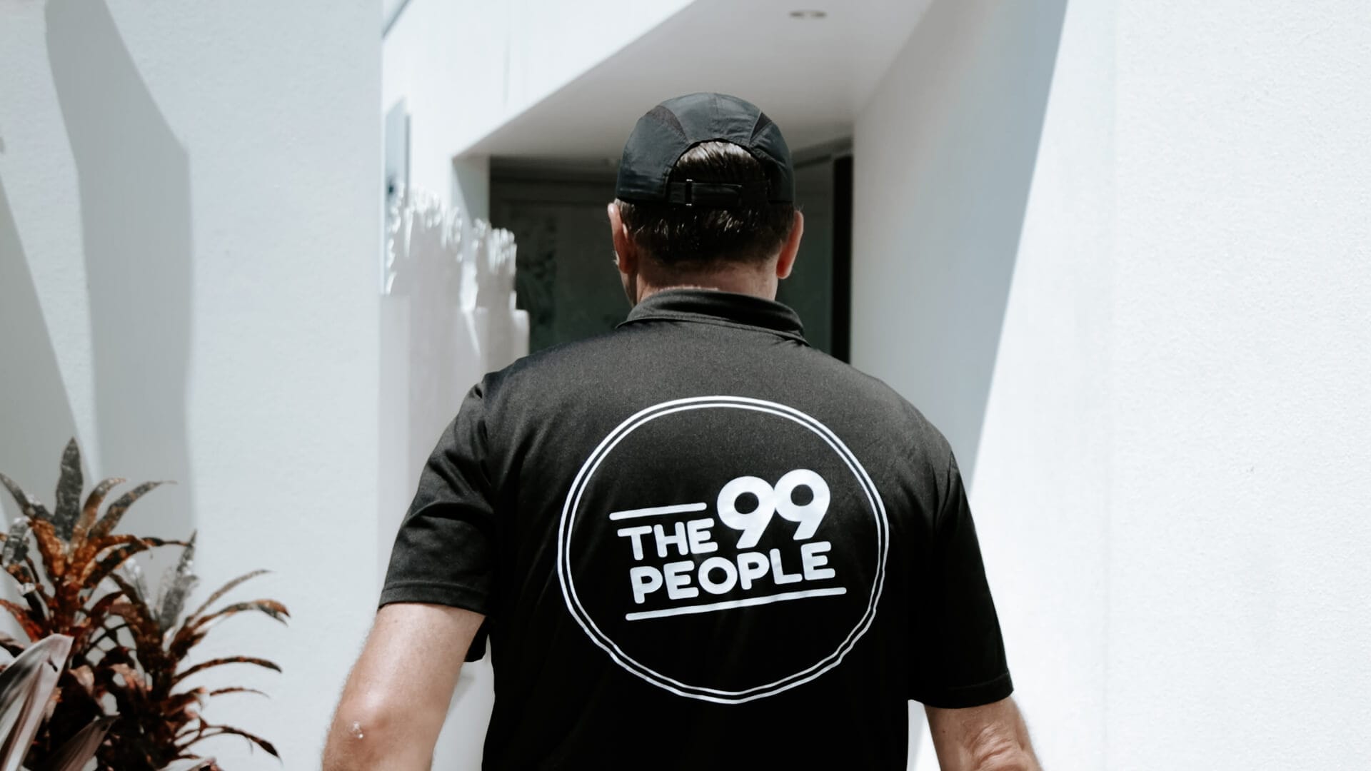 A person wearing a black cap and a black shirt with a "THE 99 PEOPLE" logo walks toward a white building entrance in Brisbane.