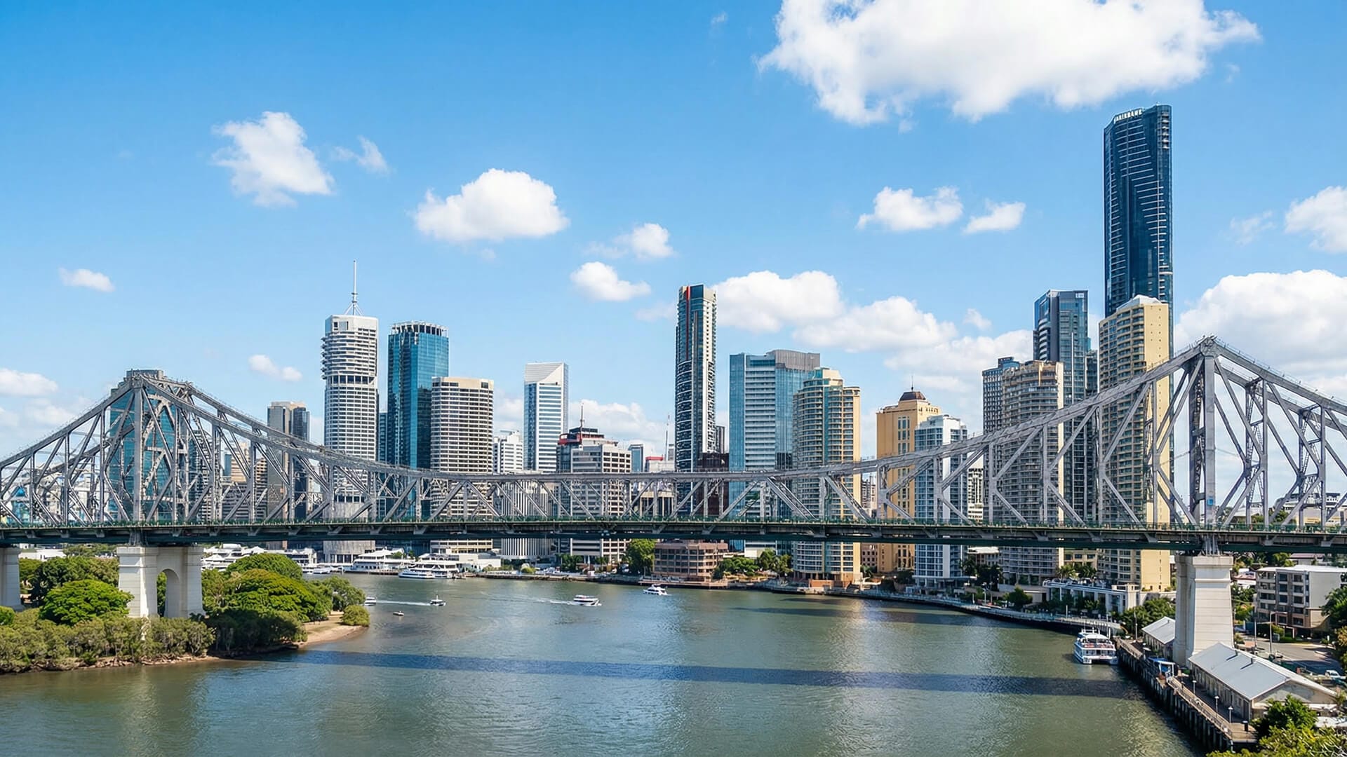 A large steel bridge spans a river with boats below, set against Brisbane's modern city skyline featuring high-rise buildings under a blue sky with scattered clouds.