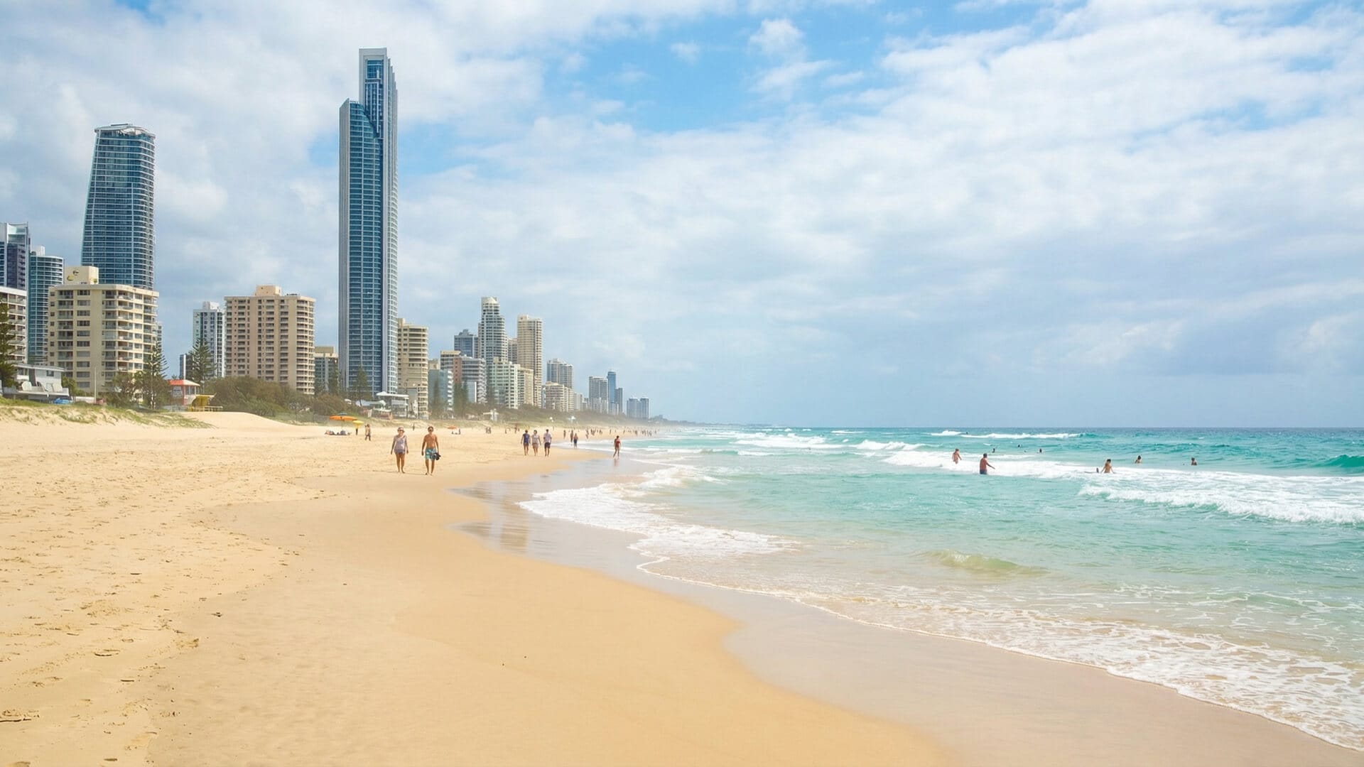 Several people walk and swim on a sandy Gold Coast beach with clear blue water; modern high-rise buildings line the coast under a partly cloudy sky.