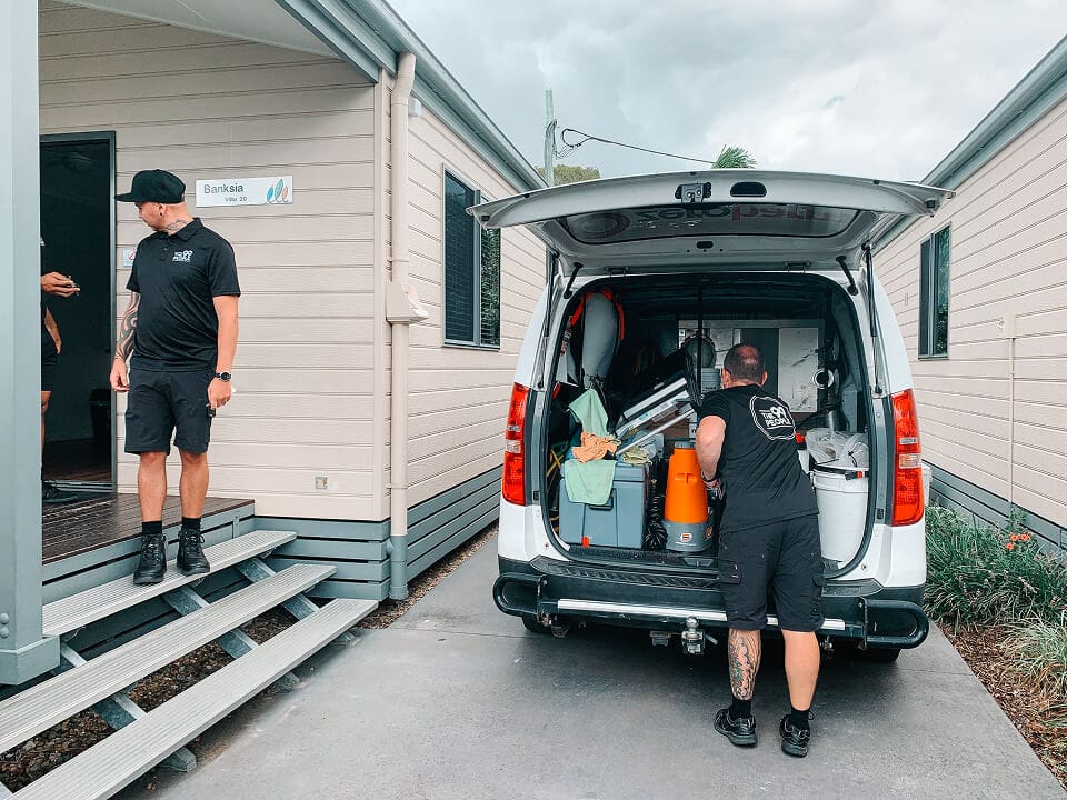 Two people in black uniforms unload cleaning equipment from the back of a white van parked beside a building labeled "Banksia.