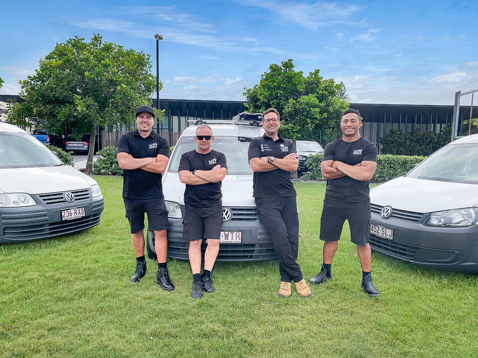 Four men in matching black uniforms stand with arms crossed in front of three white vans on a grassy area, with trees and a building in the background under a blue sky.