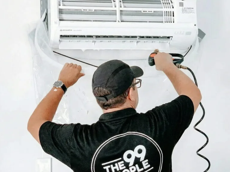 A person in a black shirt and cap is cleaning an air conditioner mounted on a wall using a spray tool.