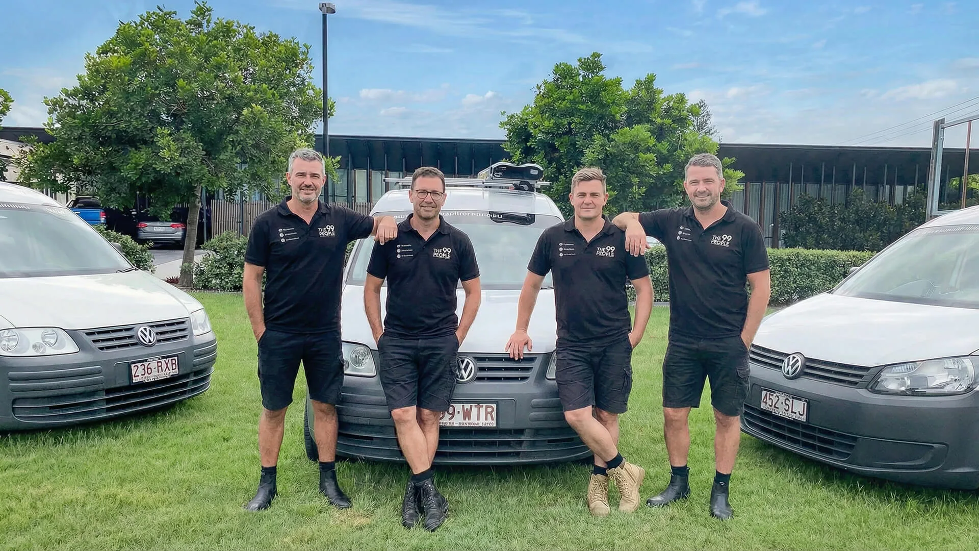 Four men in matching black uniforms stand in front of a white van, ready for Air Conditioner Cleaning Brisbane, with two other vans parked on either side on a grassy area surrounded by trees and a building in the background.