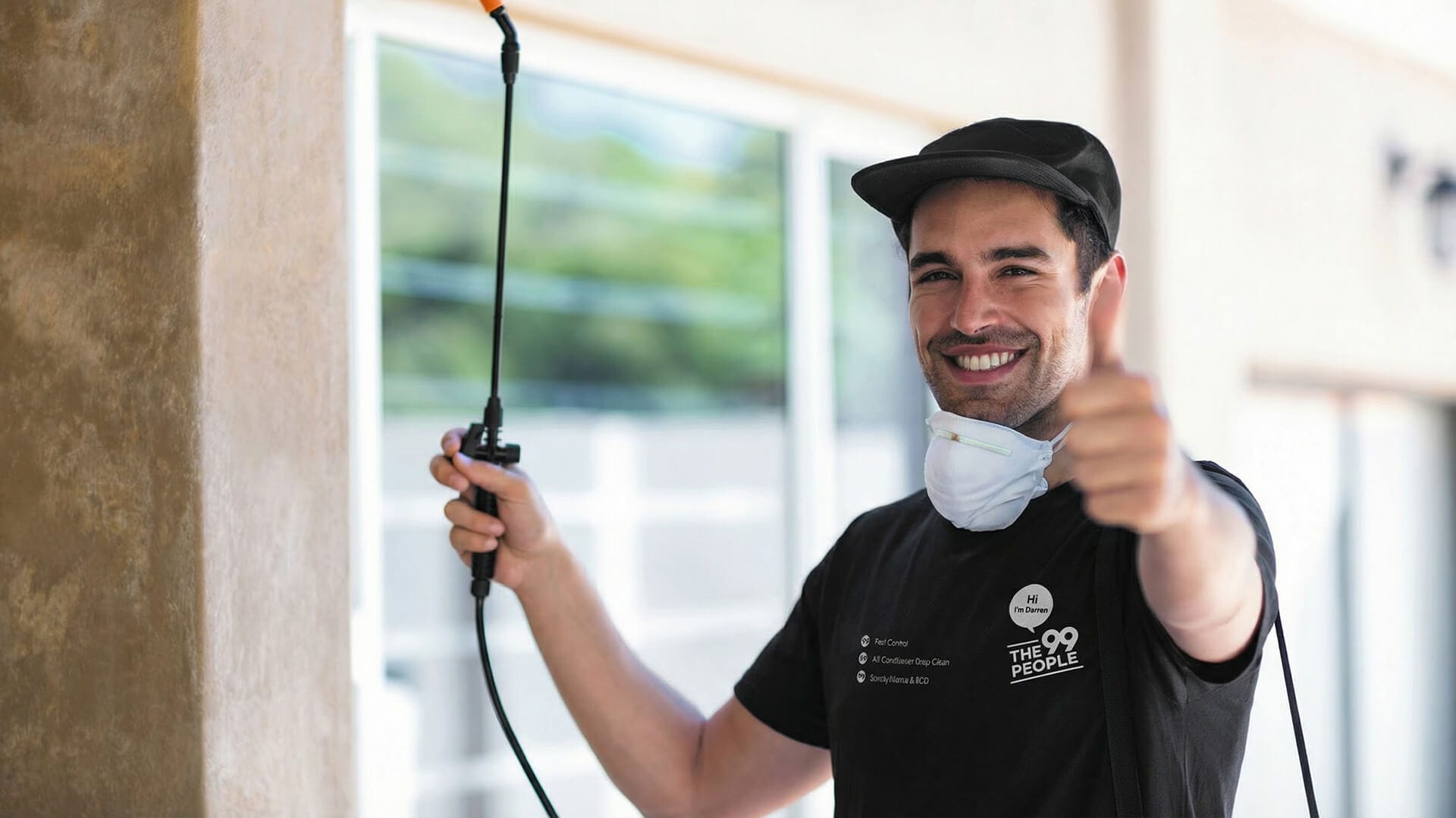 A man wearing a cap, t-shirt, and face mask around his neck holds a ant pest control spray wand for ant pest control and gives a thumbs-up gesture in front of a building.