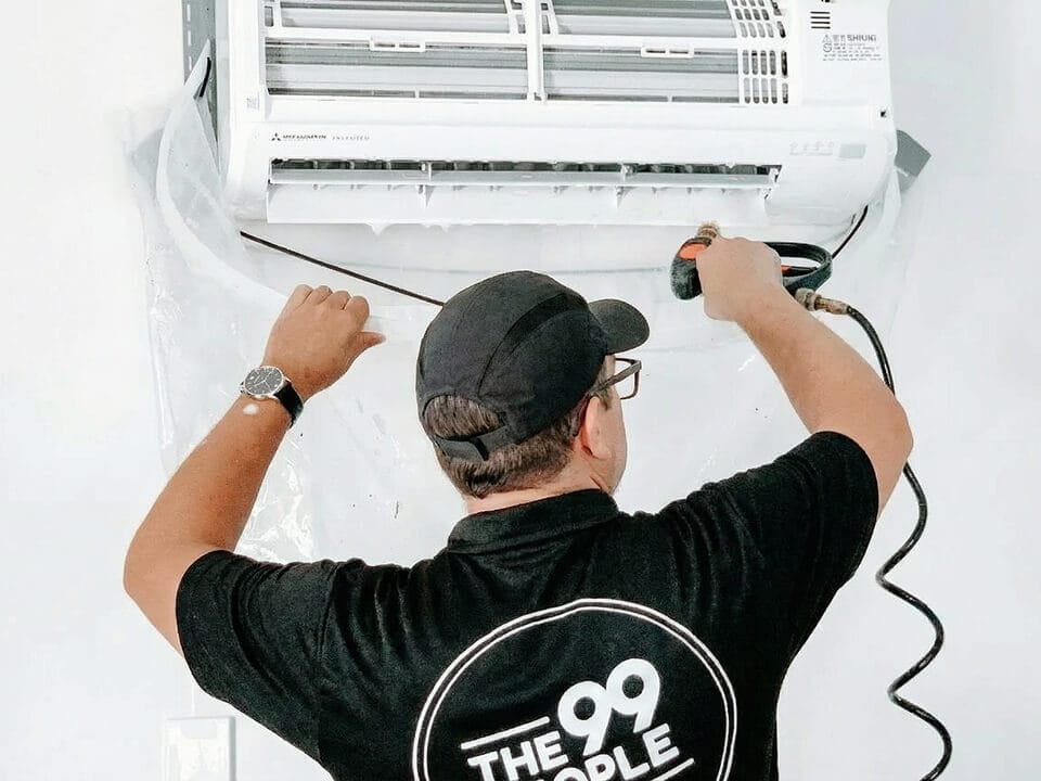 A technician in a black shirt and cap cleans a wall-mounted air conditioner with a spray tool in Brisbane.