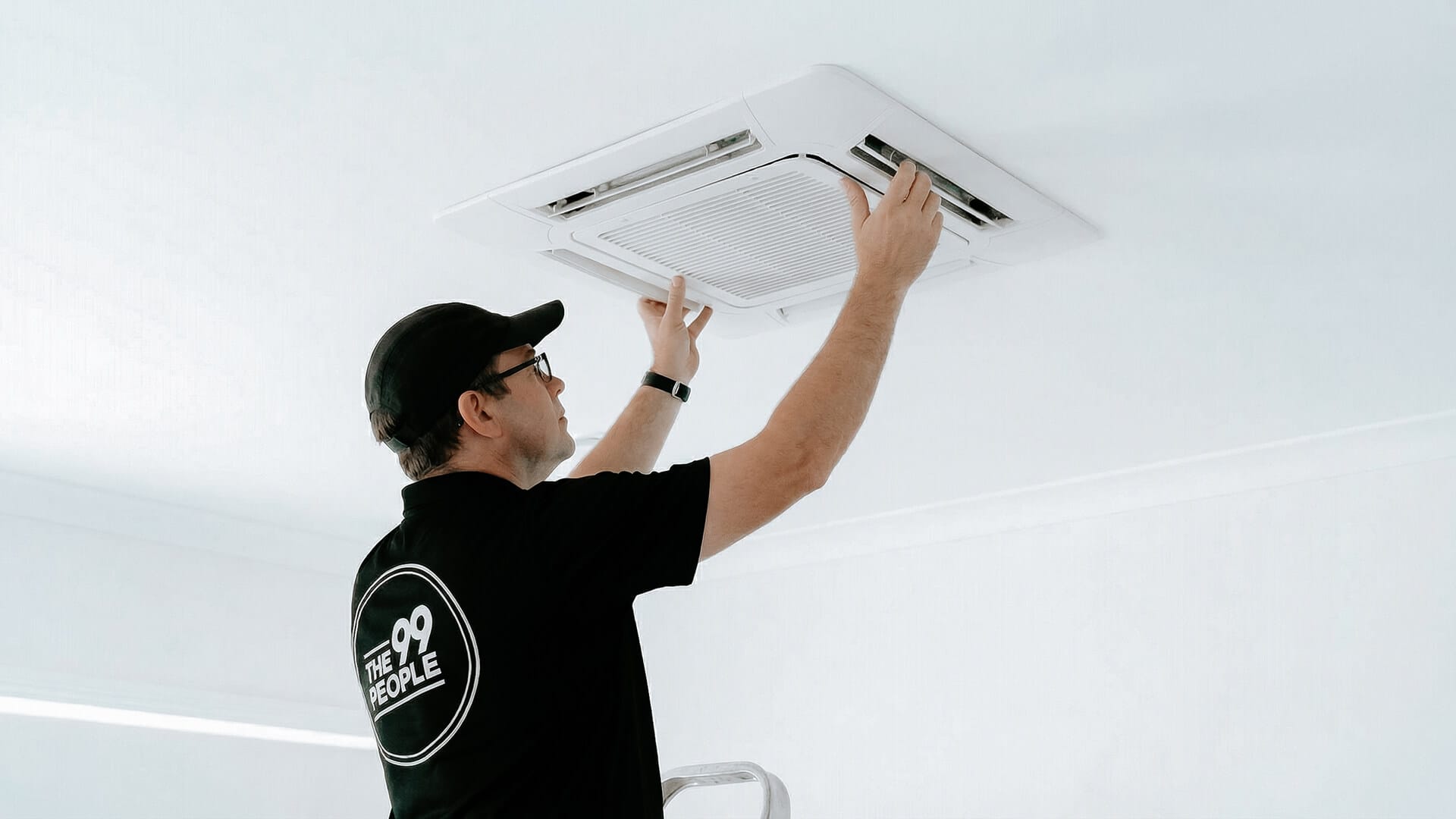 A person in a black shirt and cap is performing cassette air conditioner cleaning or servicing on a ceiling-mounted unit in a white room.