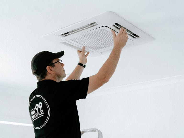 A technician in a black shirt and cap is inspecting a ceiling-mounted cassette air conditioning unit.