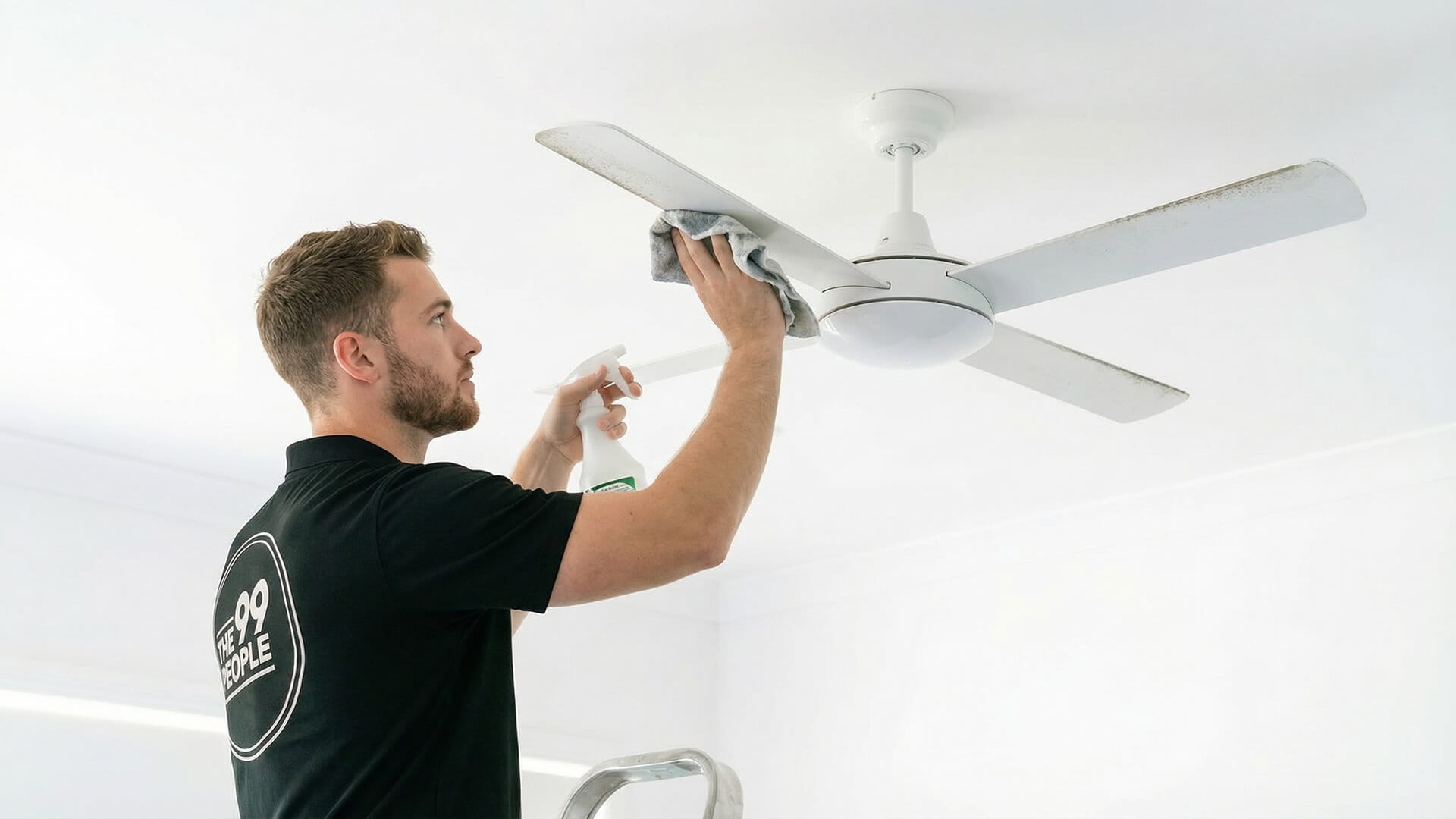 A man standing on a ladder performs ceiling fan cleaning, using a cloth and spray bottle to wipe down the white fan.