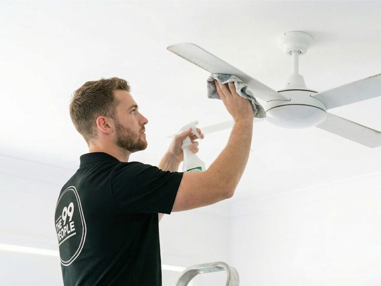 A man in a black polo shirt stands on a ladder, cleaning a white ceiling fan with a cloth and spray bottle.