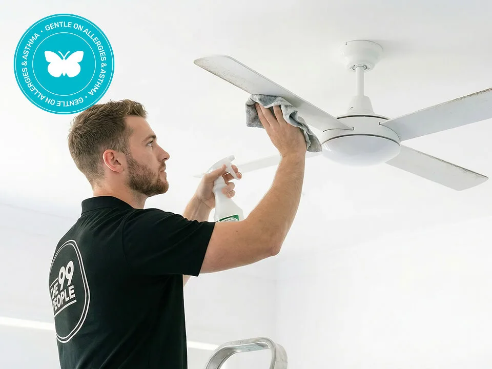 A man cleans a ceiling fan with a cloth and spray cleaner. A circular badge in the corner reads, “Gentle on Allergies & Asthma.”.