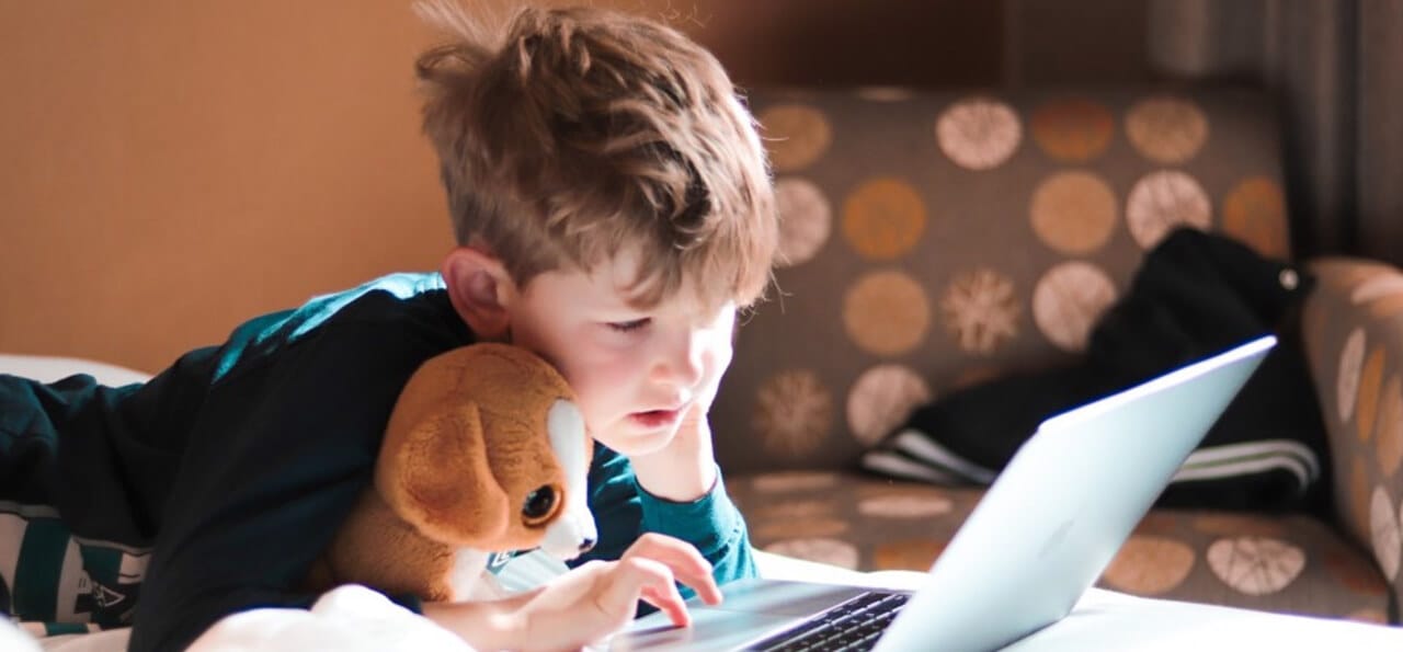 A young boy lying on a bed uses a laptop while holding a stuffed animal under his arm.