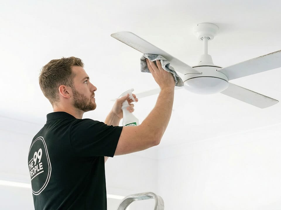 A man in a black polo shirt cleans a white ceiling fan with a cloth and spray bottle while standing on a stepladder.
