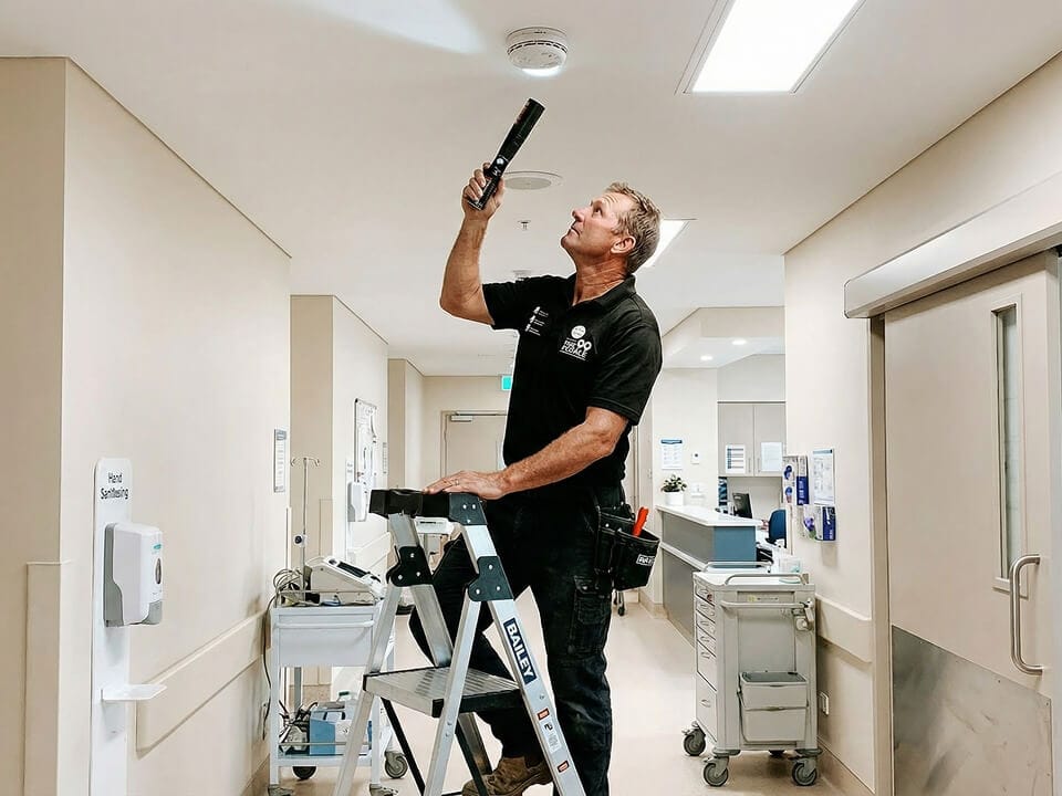 A maintenance worker standing on a ladder tests a ceiling-mounted smoke detector with a tool in a well-lit hallway.
