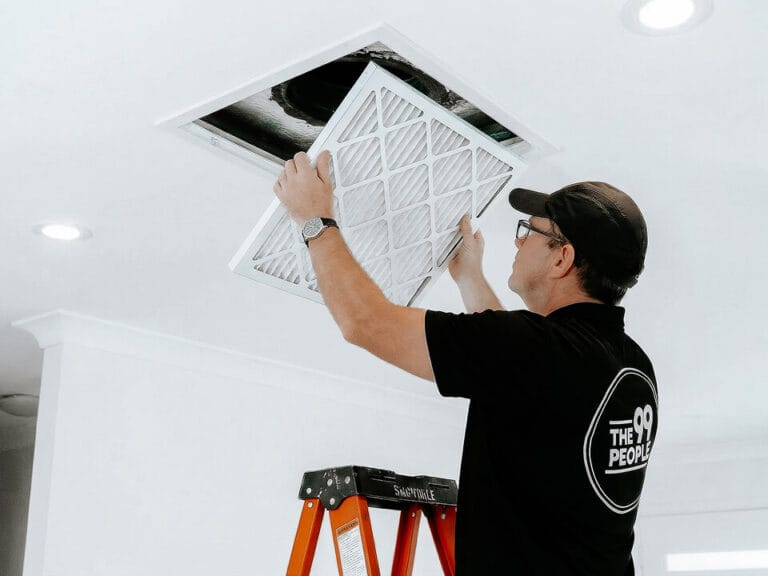 A man standing on a ladder is performing ducted air conditioner cleaning, installing or replacing an air filter in a ceiling vent.