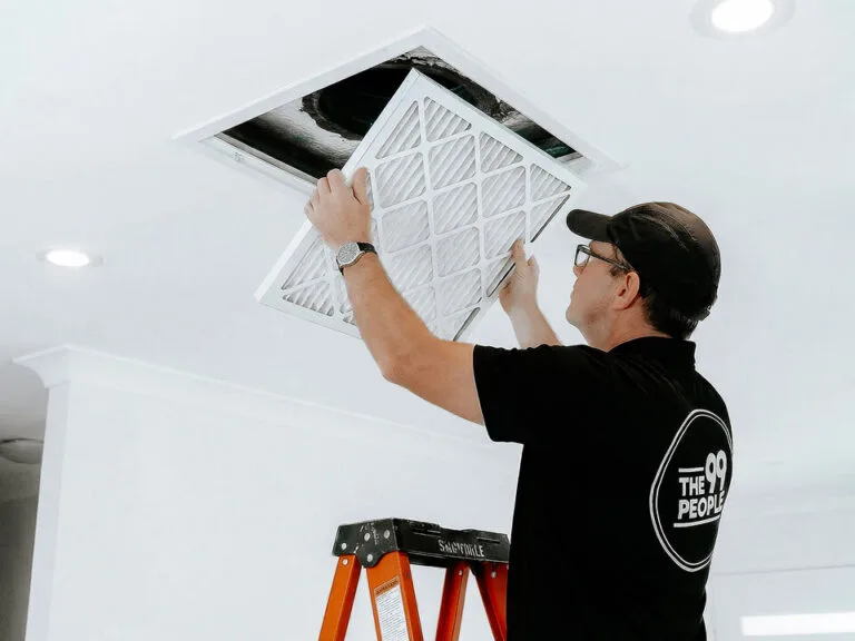 A man standing on a ladder is performing ducted air conditioner cleaning, installing or replacing an air filter in a ceiling vent.