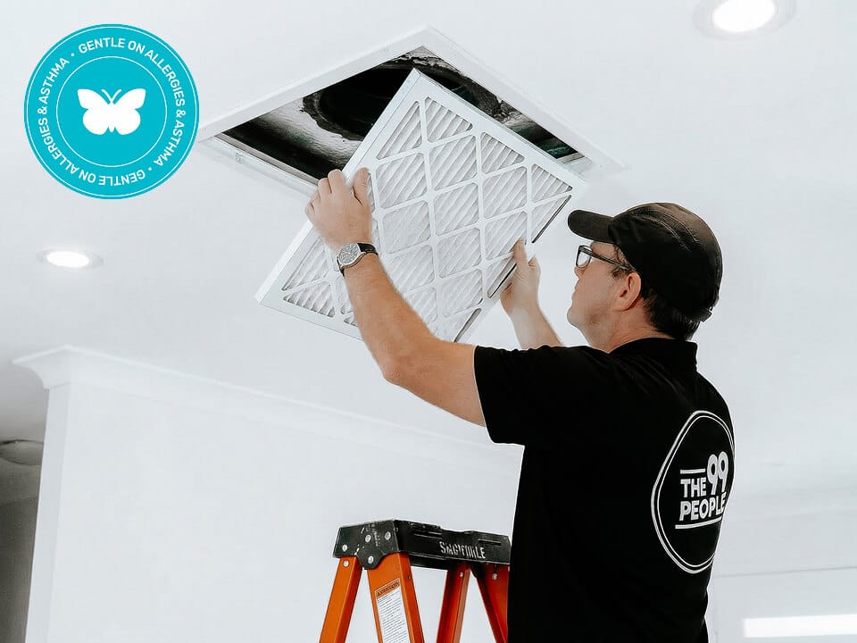 A man on a ladder replaces a ducted aircon air filter in a ceiling vent. He wears glasses, a cap, and a black shirt with a logo. A blue allergy-friendly badge appears in the top left corner.