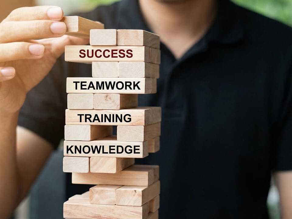 A person stacks wooden blocks labeled "KNOWLEDGE," "TRAINING," "TEAMWORK," and "SUCCESS," illustrating a progression toward success.