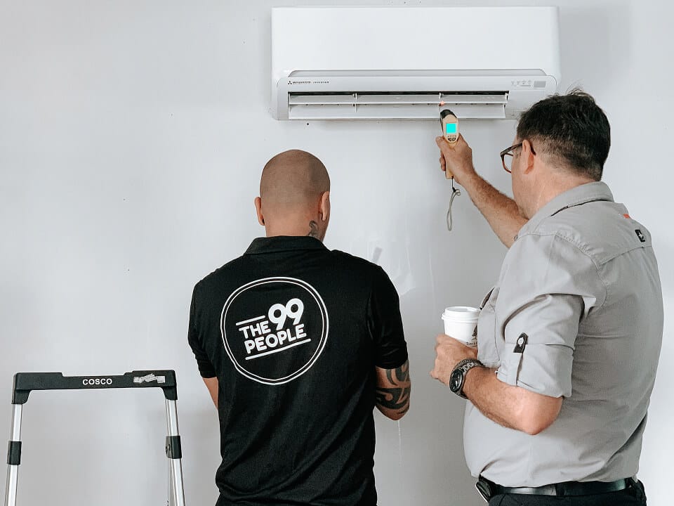 Two men inspect a wall-mounted air conditioning unit; one uses a tool while the other observes and holds a cup. A small step ladder is beside them.