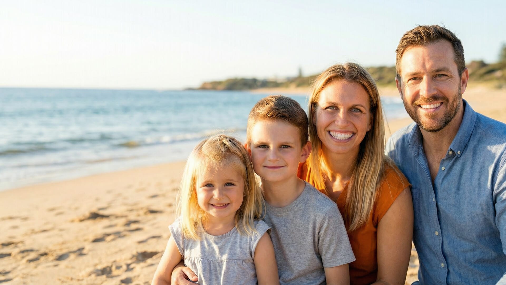 A man, woman, and two young children sit together and smile on a sandy beach with the ocean in the background.