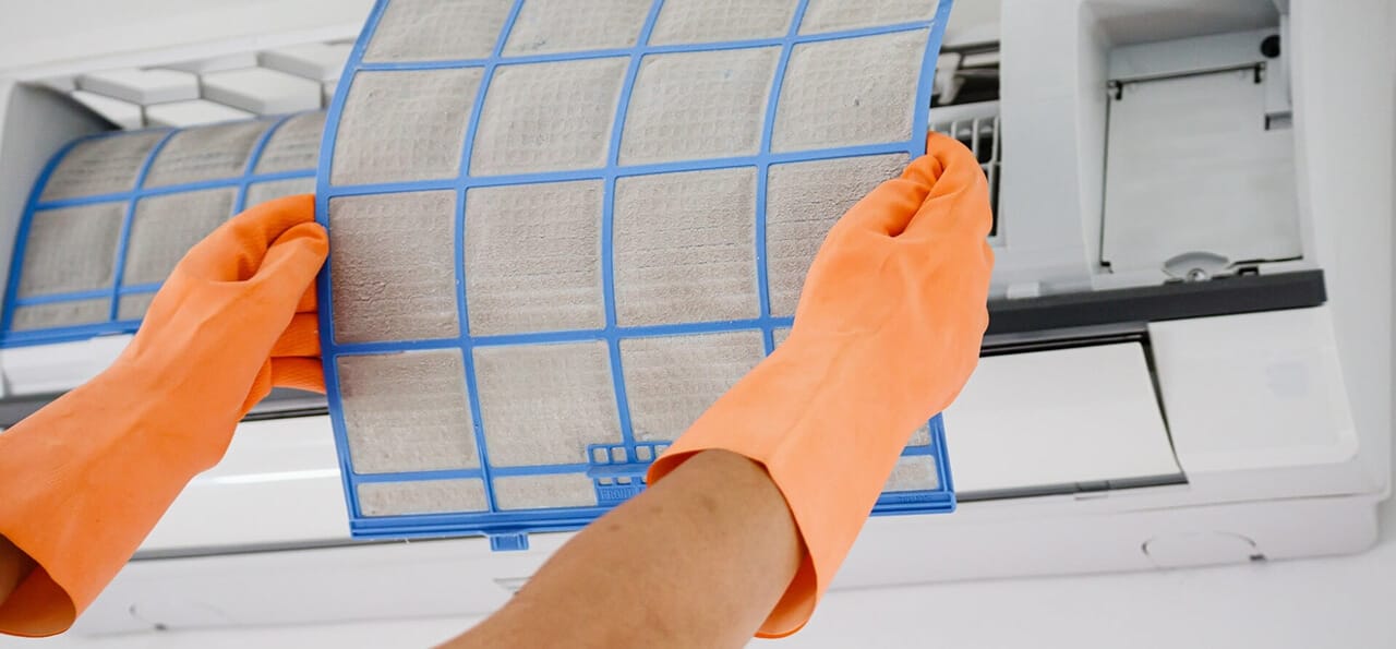 Person wearing orange gloves removes a dirty air filter from a wall-mounted air conditioning unit for cleaning or replacement.