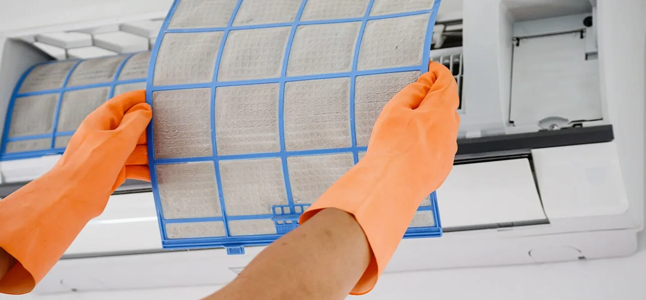 Person wearing orange gloves removes a dirty air filter from a wall-mounted air conditioning unit for cleaning or replacement.
