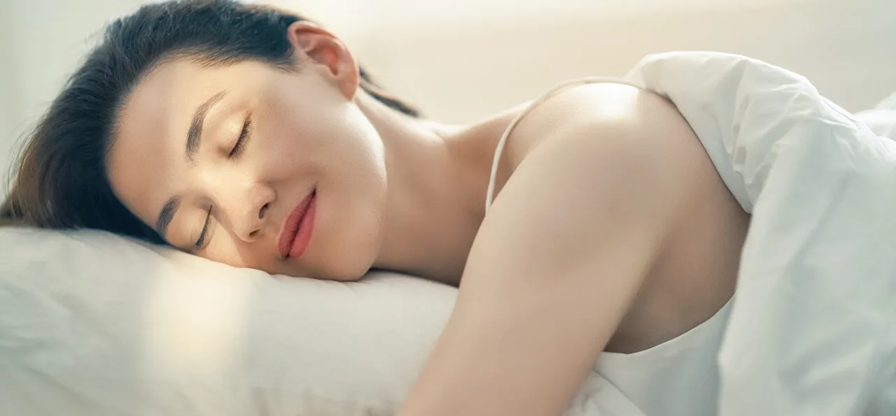 A woman with closed eyes sleeps peacefully in bed under white sheets, with soft natural light illuminating her face.