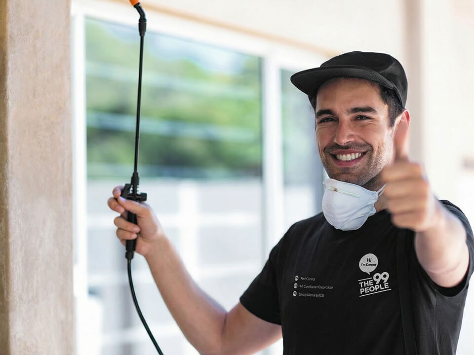 A man wearing a cap and face mask around his neck holds a pest control sprayer and gives a thumbs up outside a building in Noosa.