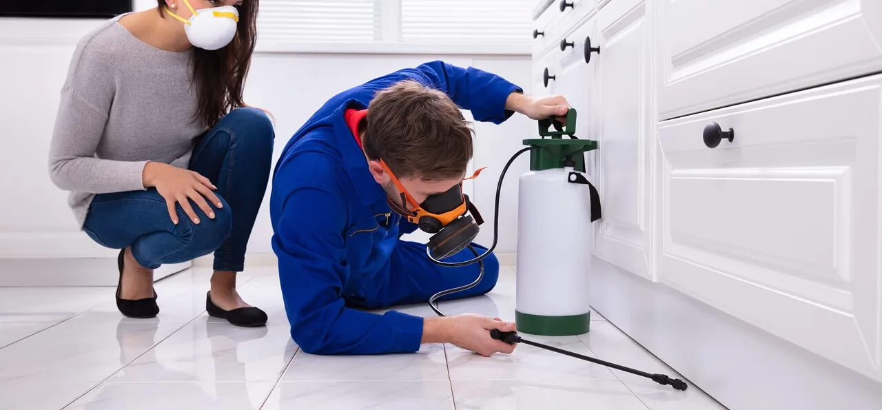 A pest control worker in protective gear sprays insecticide along a kitchen baseboard while a woman wearing a mask watches nearby.
