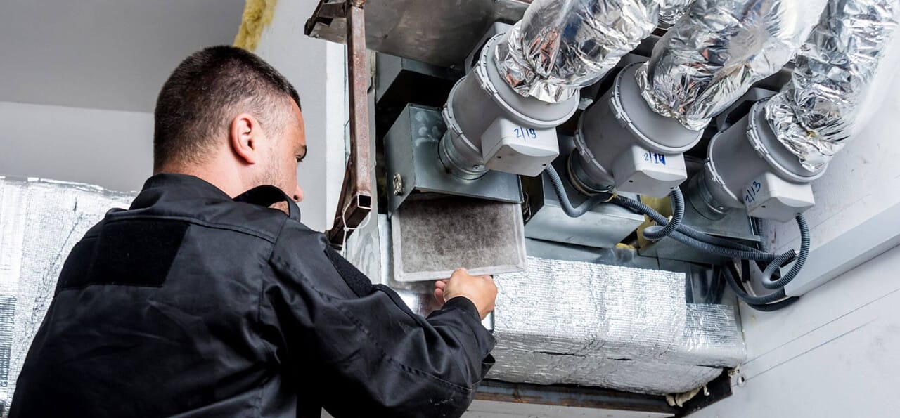 A technician in black workwear inspects and replaces a dirty air filter in an HVAC ventilation system with insulated ducts.