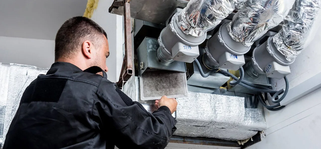 A technician in black workwear inspects and replaces a dirty air filter in an HVAC ventilation system with insulated ducts.