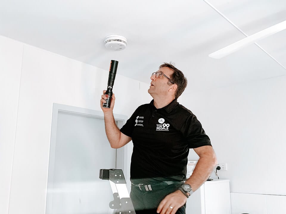 A man in a black shirt stands on a ladder, holding a tool to test a smoke detector on the ceiling in a white Gold Coast room.