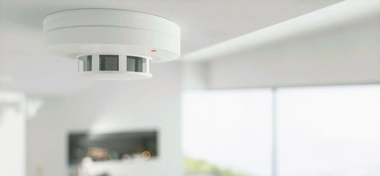 A white smoke detector is mounted on a white ceiling in a bright, modern room with blurred windows in the background.