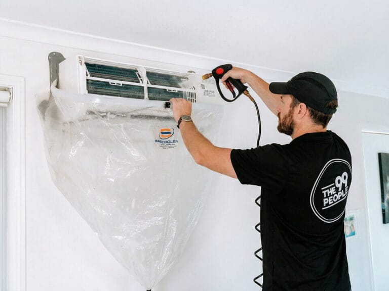 A technician cleans a wall-mounted split air conditioner using professional equipment, with a protective plastic cover to catch debris.
