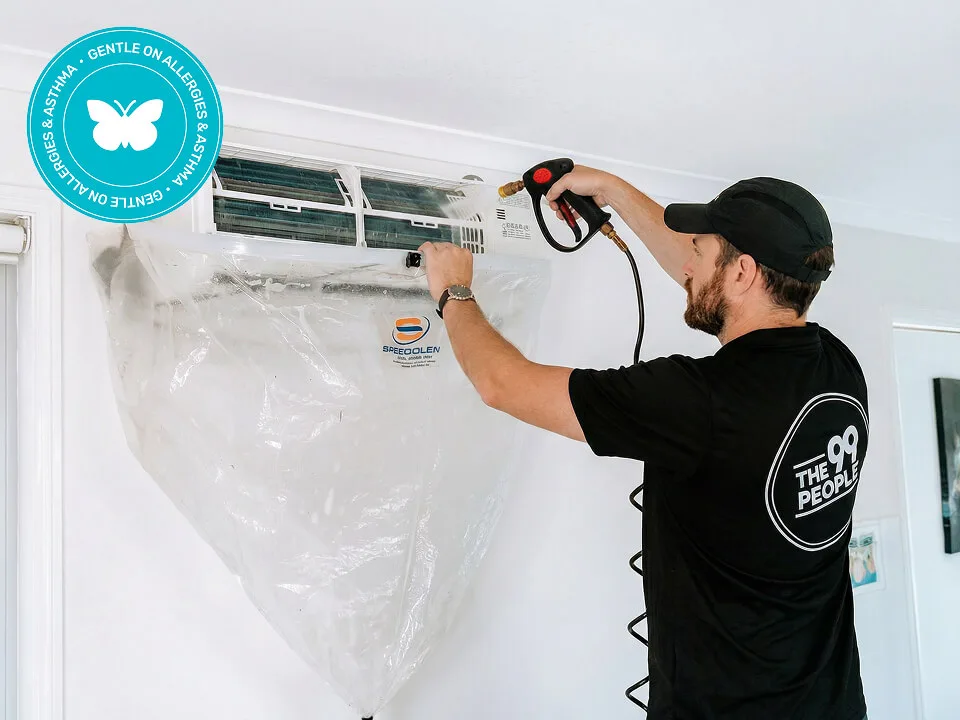 A technician cleans a wall-mounted split air conditioner using a spray tool, with a protective plastic cover underneath to catch debris.