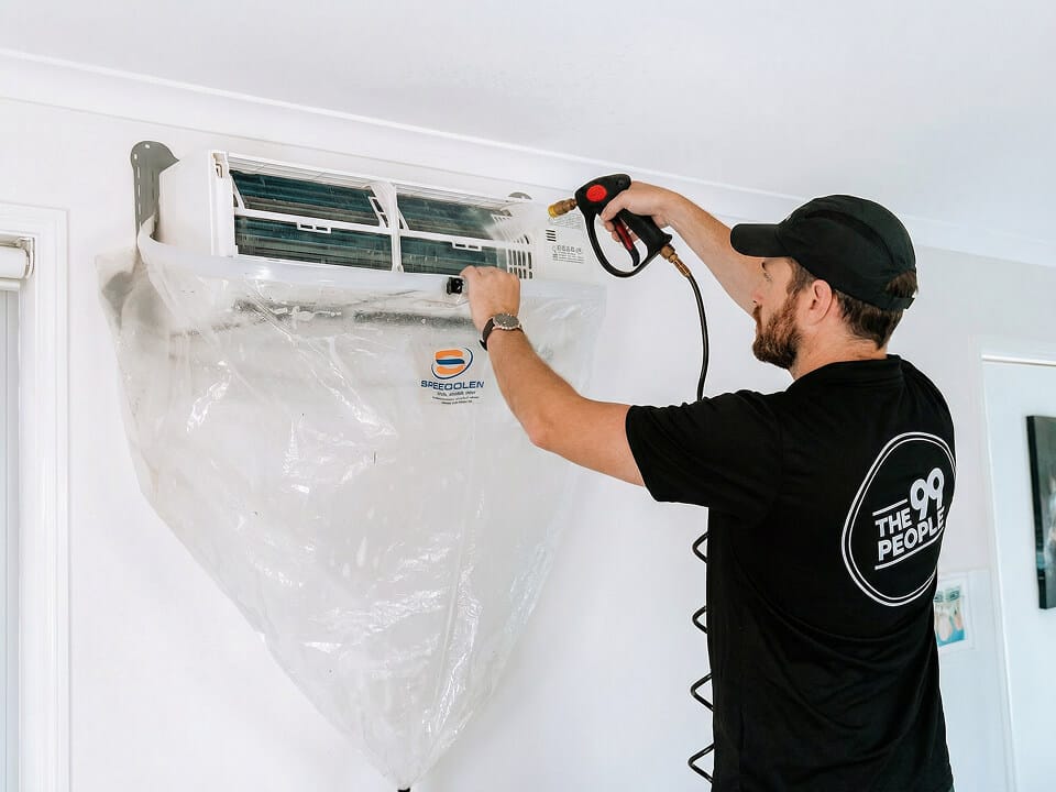 A technician cleans a wall-mounted split air conditioner using professional equipment, with a protective plastic cover to catch debris.