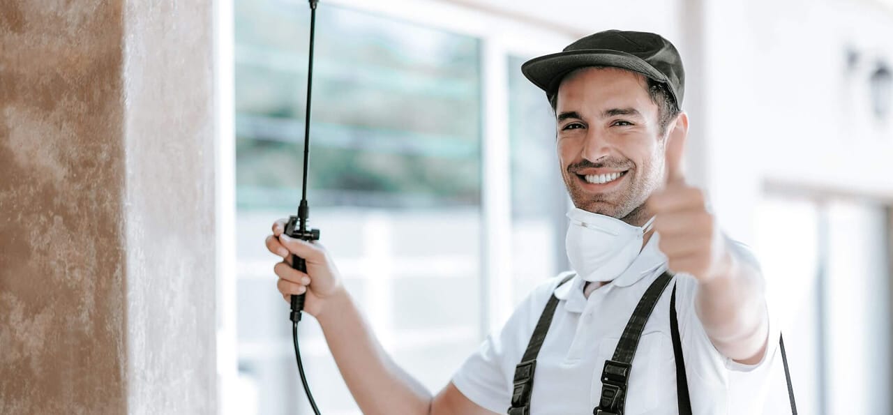 A man wearing a cap, mask, and overalls holds a spraying tool and gives a thumbs-up gesture, standing indoors near a window.
