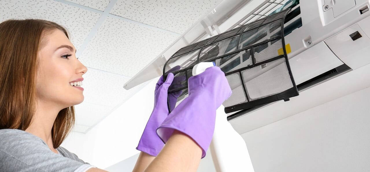 Woman wearing purple gloves sprays and cleans an air conditioner filter indoors.