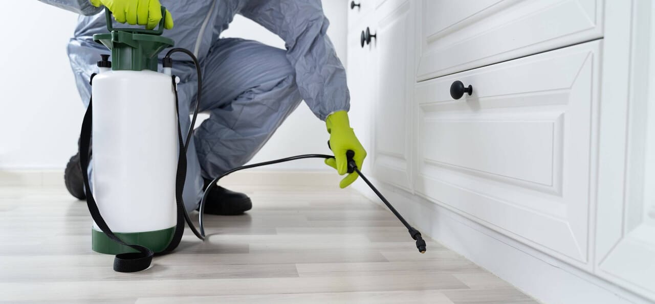 Person wearing gloves and protective suit sprays pesticide along the baseboard of a kitchen cabinet with a handheld sprayer.