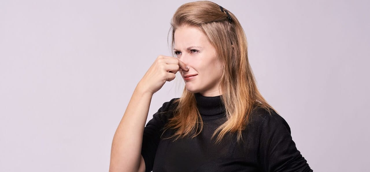 A woman with long blonde hair wearing a black top is holding her nose and appears to be reacting to an unpleasant smell.