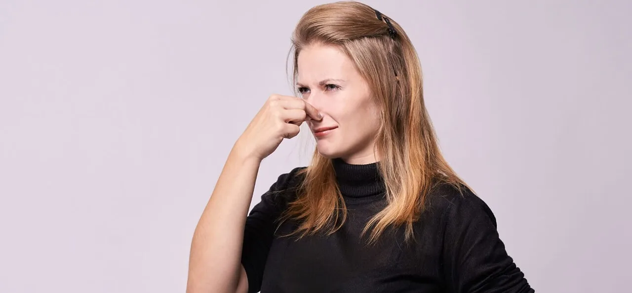 A woman with long blonde hair wearing a black top is holding her nose and appears to be reacting to an unpleasant smell.